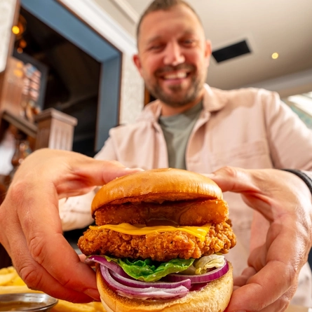 A close up image of a burger with a person in the background within the interior restaurant and seating area at a Hungry Horse venue.