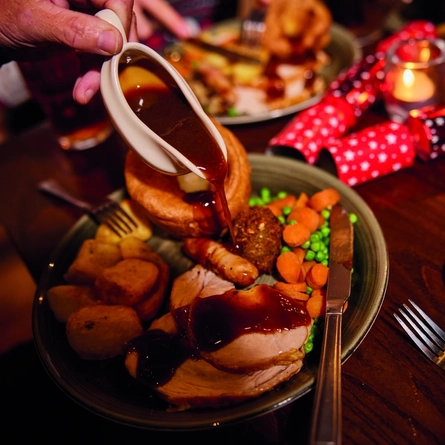 A hand poring gravy from a gravy boat onto a plated roast dinner in a festive setting.