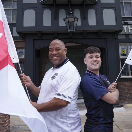 John Barnes and Mark Thorburn standing back to back, outside in front of The Endbutt in Liverpool. John is wearing an England football shirt and holding the English flag, Mark is wearing a Scotland shirt and holding the Scottish flag.