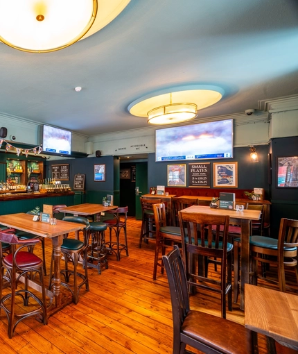 The bar and interior restaurant seating area at the Royal George in Euston; with wooden flooring, an upholstered booth seat, TVs on the walls, and gaming machines.