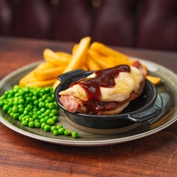 A roasting dish containing Hunters Chicken with plated chips and Peas on a table.