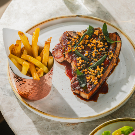 An advertising image showing a plated Dry Aged Pork T-Bone dish and a pint of ale sat on a table within the exterior beer garden seating area at a Crafted venue.