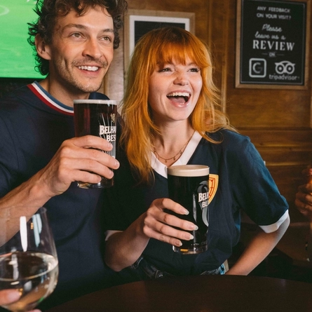 An image focusing on 2 people within a group of friends stood at a table watching a football match and enjoying a selection of drinks and snacks within the interior seating area at a Greene King venue.