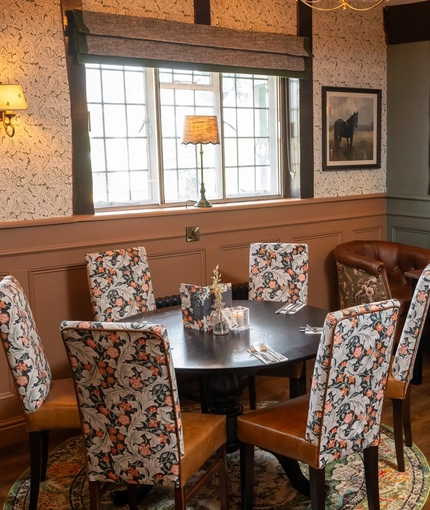 The interior restaurant seating area at The Hollybush in Lichfield, with upholstered chairs, bottles of wine in a glass fronted cabinet, a lamp on the windowsill, and a large potted plant.