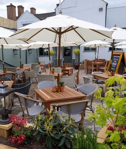 The exterior beer garden seating area of The Hollybush in Lichfield, with shade umbrellas and potted plants on each of the wooden tables, and wooden planter boxes containing plants, flowers, and lanterns.