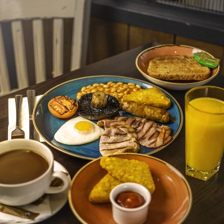 A image of a served full English breakfast with a side of hash browns and toast, also a orange juice and cup of tea, served at Flaming Grill venues.