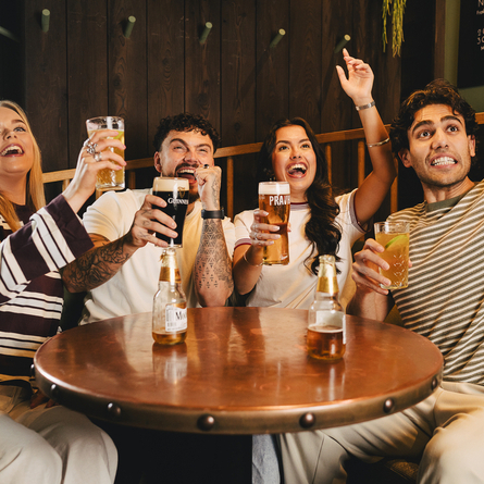 A group of friends sat round a table in the interior seating area in a Crafted venue with drinks at the table, the group are all cheering with their hands in the air.