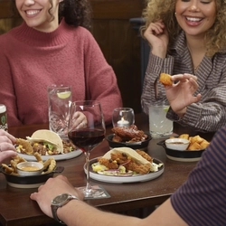 A group of people sitting around a wooden table at a restaurant, sharing several small plates of food including tacos, fried items, and dipping sauces. Drinks are on the table. The individuals are engaged in conversation while eating, with one person holding a piece of food and others seated around the table.