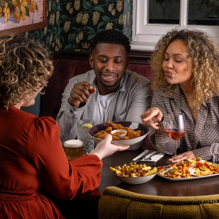 Three people sit around a wooden table in a restaurant booth. One person seated on the far side of the table is holding a piece of fried food toward the person sitting beside them. A third person, is seated facing the pair and reaching forward with a large bowl filled with shared appetizers. Other shared dishes are spread across the table.
