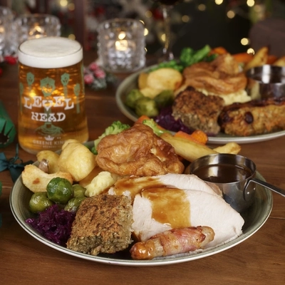 A close up view of a Roast Turkey dinner, featuring slices of roast turkey, a Yorkshire pudding, roast potatoes, vegetables, pigs in blankets, and gravy, served on a plate. The plate sits on a table with a glass of Level Head IPA, another plate of food, and Christmas table decorations. 