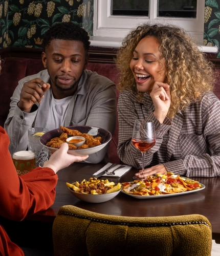 Three people sit around a wooden table in a restaurant booth. One person seated on the far side of the table is holding a piece of fried food toward the person sitting beside them. A third person, is seated facing the pair and reaching forward with a large bowl filled with shared appetizers. Other shared dishes are spread across the table.
