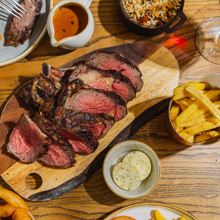 An advertising image showing a boarded Ribeye Sharer dish including onions rings, chips, salad and sauce sat on a table within the interior restaurant seating area at The Four Oaks.