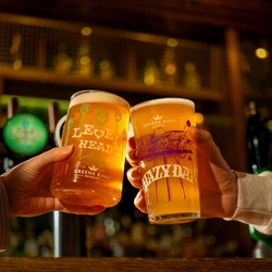 A close up view of two hands clinking together a glass of Level Head IPA and a glass of Hazy Day IPA, at the bar inside a pub.