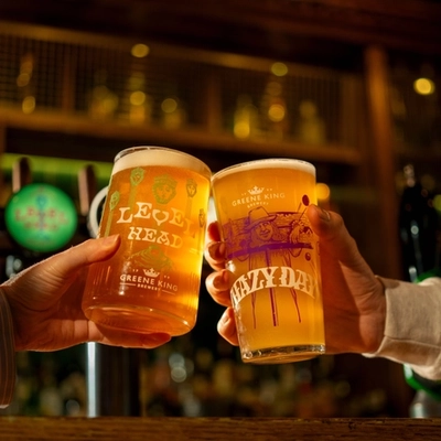 A close up view of two hands clinking together a glass of Level Head IPA and a glass of Hazy Day IPA, at the bar inside a pub.