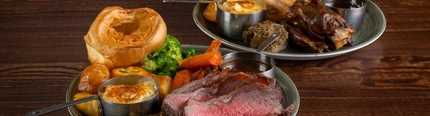 Two plates of roast dinner, including meat, Yorkshire pudding, vegetables and gravy, sitting on a wooden table.