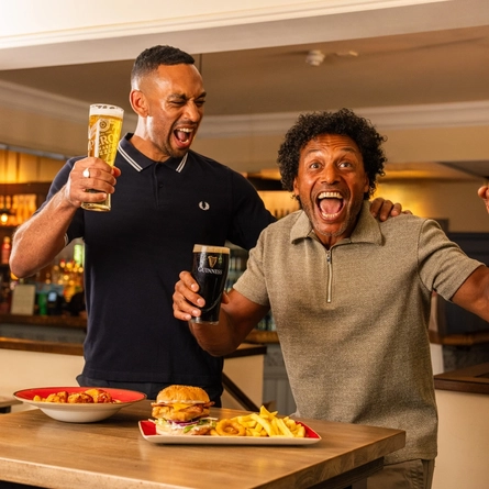 Two people standing beside a high table in a pub, holding a pint of lager and a pint of Guinness. Plates of food, including a burger with chips and a pasta dish, are on the table. The pub interior features a stocked bar, exposed brickwork and warm lighting in the background.