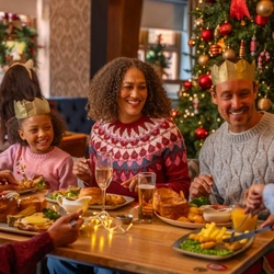 A family enjoys a festive Christmas meal at a dining table beside a decorated Christmas tree adorned with red and gold baubles, candy cane ornaments and warm fairy lights. Several diners wear paper crowns from Christmas crackers while sharing food and conversation. The table is set with traditional roast dinners, gravy boats, glasses of orange juice, sparkling drinks and Christmas crackers. Seasonal decorations and warm lighting create a cosy Christmas restaurant setting.