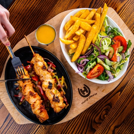 Hands using a knife and fork to cut into chicken skewers which are served on a plate next to another plate of chips and salad.