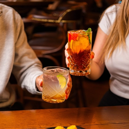 A close up view of two people at a wooden table in a pub, holding cocktails made with Pimms and Margarita.
