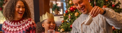 A festive Christmas meal taking place beside a decorated Christmas tree adorned with red and gold baubles, candy cane ornaments and warm fairy lights. Three people seated at a dining table wear paper crowns from Christmas crackers while enjoying a traditional festive meal. One person pours gravy from a gravy boat onto a dinner plate. The table is set with roast dinners, vegetables, glasses of orange juice, a pint of beer, Christmas crackers and string lights, creating a warm and seasonal restaurant setting.