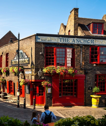 The exterior facade, signage, and seating area of The Anchor Bankside in Southwark; with bright red window frames; hanging flower baskets on the brick walls; and flower boxes below the upper window sills.