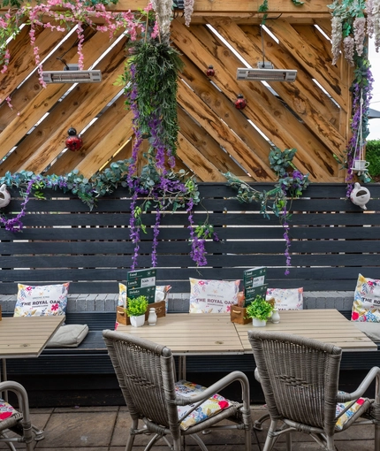 An exterior beer garden seating area at The Royal Oak, with wicker chairs and bench seating underneath a wooden pergola decorated with artificial flowers.