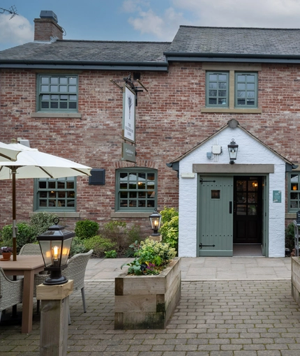 The exterior facade, main entrance, and beer garden seating area at The Malthouse Farm, with wicker chairs, wooden planter boxes, and shade umbrellas.