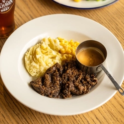 Haggis, Neeps and Tatties served on a plate with a small pot of sauce. The plate sits on a wooden table next to a glass of Belhaven Best ale and another plate of food.