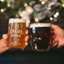 A close up view of two hands; one of them holding a glass of Level Head, the other holding a glass tankard of beer. A Christmas garland lit with fairy lights is visible in the background.