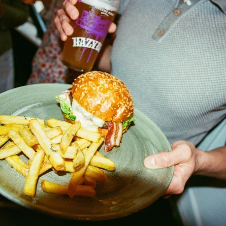 A main of burger and chips served on a plate being held by a customer and a pint of Hazy Day in the other hand in a Greene King London venue.