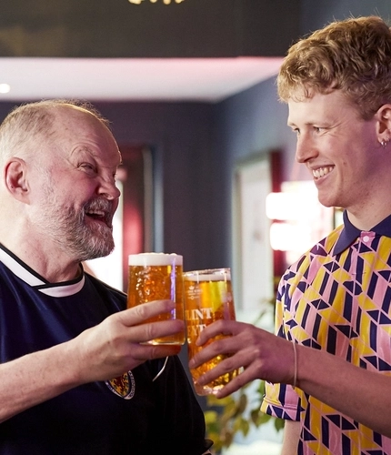 A group of people standing around a table in a pub; two of them are laughing and clinking together their glasses of beer.