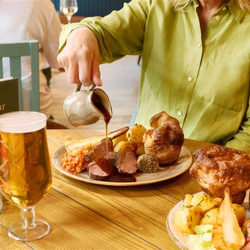 An image showing a person pouring gravy over a Sunday Roast plated dish sat at a table within the interior restaurant seating area at a Chef & Brewer venue.