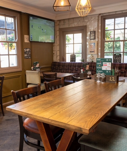 An interior restaurant seating area at The Woolpack Inn; with wooden flooring, an upholstered booth seat against the windows, and a large TV on the wall.