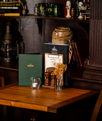 A close up view of menus, salt and pepper mills, a tealight, and a small glass vase filled with dried flowers; on a wooden restaurant table inside The London Apprentice. Books and ornaments stand on wooden shelves next to the table.