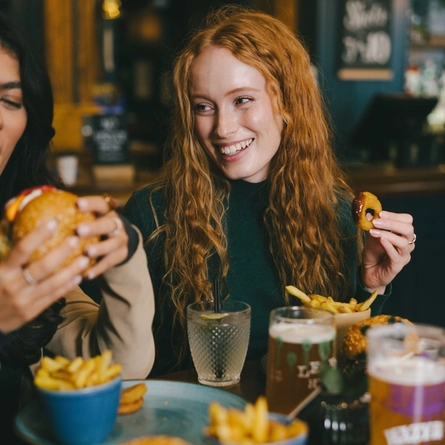 A lifestyle image of 3 friends sat within the interior restaurant seating area enjoying Burger dishes and various drinks available at Urban Core venues.