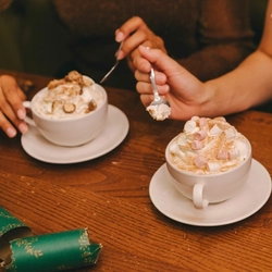 A close up image of 2 friends enjoying cups of hot chocolate with cream and marshmallows within the interior seating area at a Greene King venue.