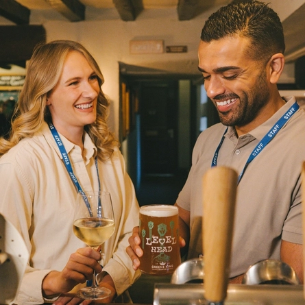 Two customers stood at the bar with a glass of white wine and a pint of Level Head at a Greene King venue.