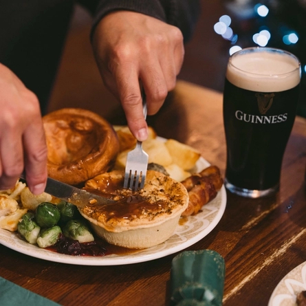 Person cutting into a festive pie served with a Yorkshire pudding, roast potatoes, pigs in blankets, Brussels sprouts and red cabbage on a wooden table. A glass and can of Guinness 0.0 alcohol-free stout sit beside the meal, with green Christmas crackers nearby.
