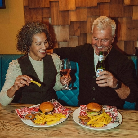An image showing 2 people sat at a table enjoying mains burger dishes and drinks at a Flaming Grill venue.