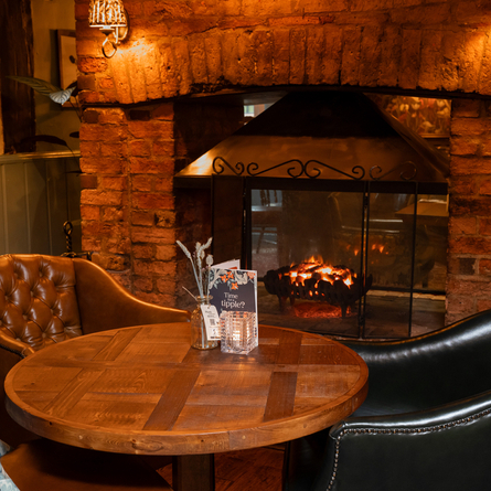 A close up view of an interior restaurant seating area at Ye Olde Swan, featuring a round wooden table, three upholstered chairs, and a double sided fireplace.