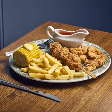 A Chick Mix meal featuring a chicken skewer, corn on the cob, fries, and a jug of dipping sauce, served on a plate which sits on a wooden table with cutlery and a glass of soft drink.