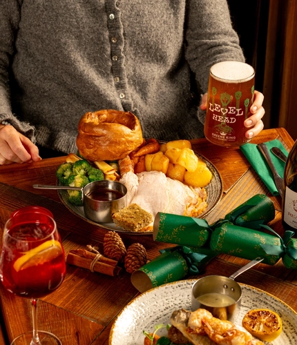 A person is seated at a wooden table. The table is decorated with festive decoration and is  holding a plated turkey dinner.  Only the torso and arm is visible in the image. The person is holding a pint of Level Head.