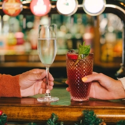 A glass of champagne and a cocktail served in a highball glass and garnished with mint, sitting on a bar inside a pub. A hand holds each glass and a Christmas garland is attached to the bar.