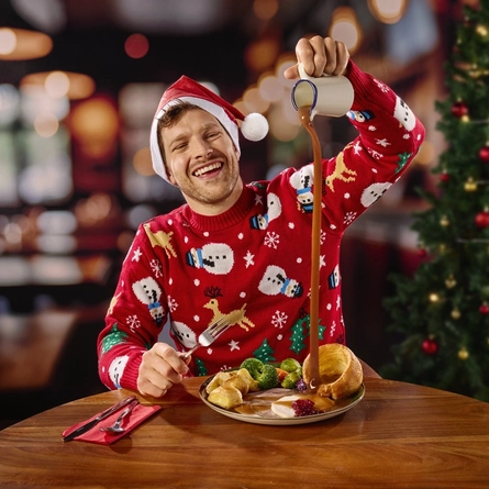 A person in a Santa hat and Christmas jumper, sitting at a wooden restaurant table and pouring gravy from a jug onto the plate of Christmas dinner in front of them. Cutlery and a red serviette also sit on the table, and a Christmas tree is visible in the background.
