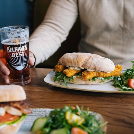 A close up image image focusing on the products with a person sat at a table within the interior restaurant seating area enjoying a drink and a sandwich dish at a Heritage venue.