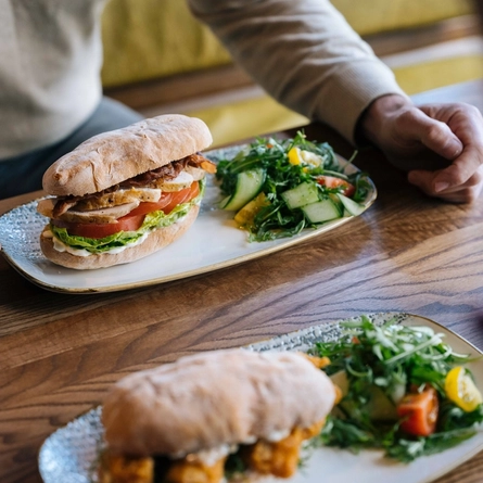 A close up image image focusing on the products with a person sat at a table within the interior restaurant seating area enjoying a drink and a sandwich dish at a Heritage venue.