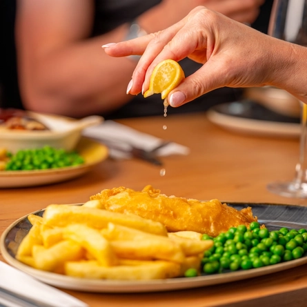 A hand squeezing a lemon wedge over a plated portion of fish and chips, at an indoor table.