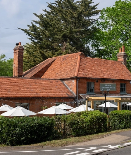 A view from across the road of the exterior facade, signage, and beer garden seating area of the Ye Olde Leathern Bottel in Barkham, with shade umbrellas and string lights. 