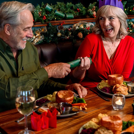 An image focusing 2 people sat within a group at a table within the interior restaurant seating area pulling a cracker while enjoying drinks and dishes from the Christmas menu at a Community Social venue.