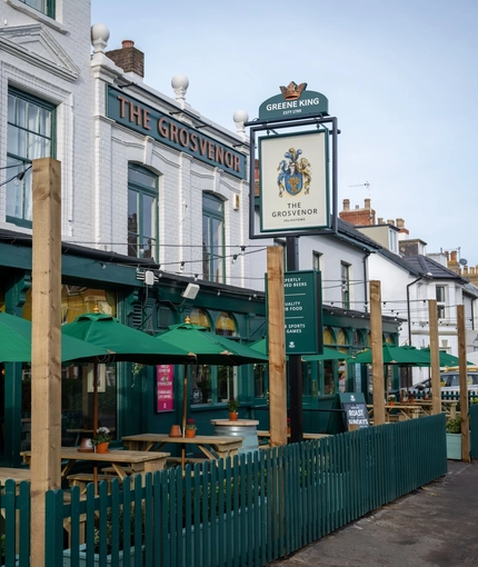 The exterior facade, signage, and seating area of The Grosvenor, with string lights above the wooden picnic tables and shade umbrellas.